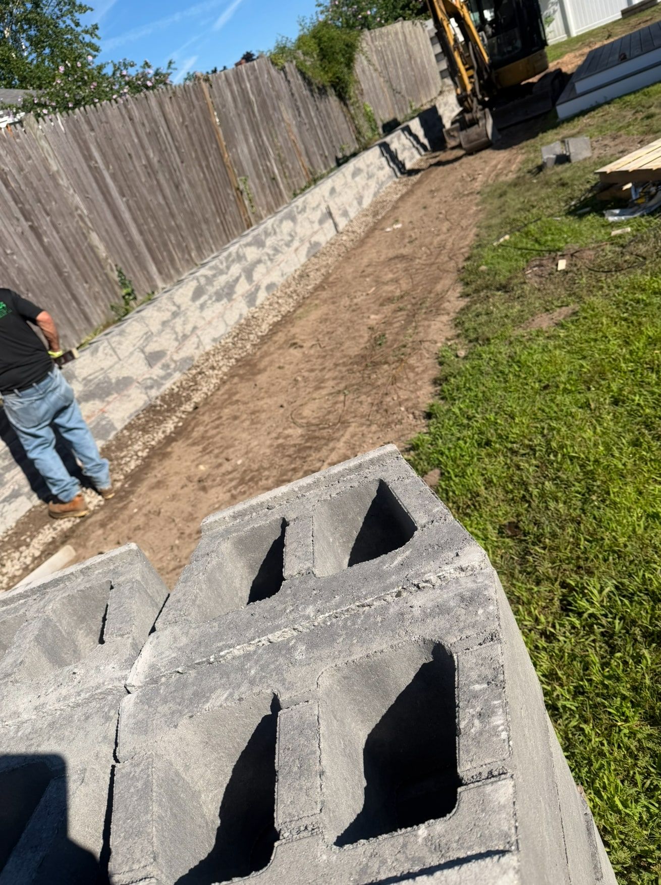 Retaining wall under construction along a fence, with a mini excavator, stacked blocks and a worker on site