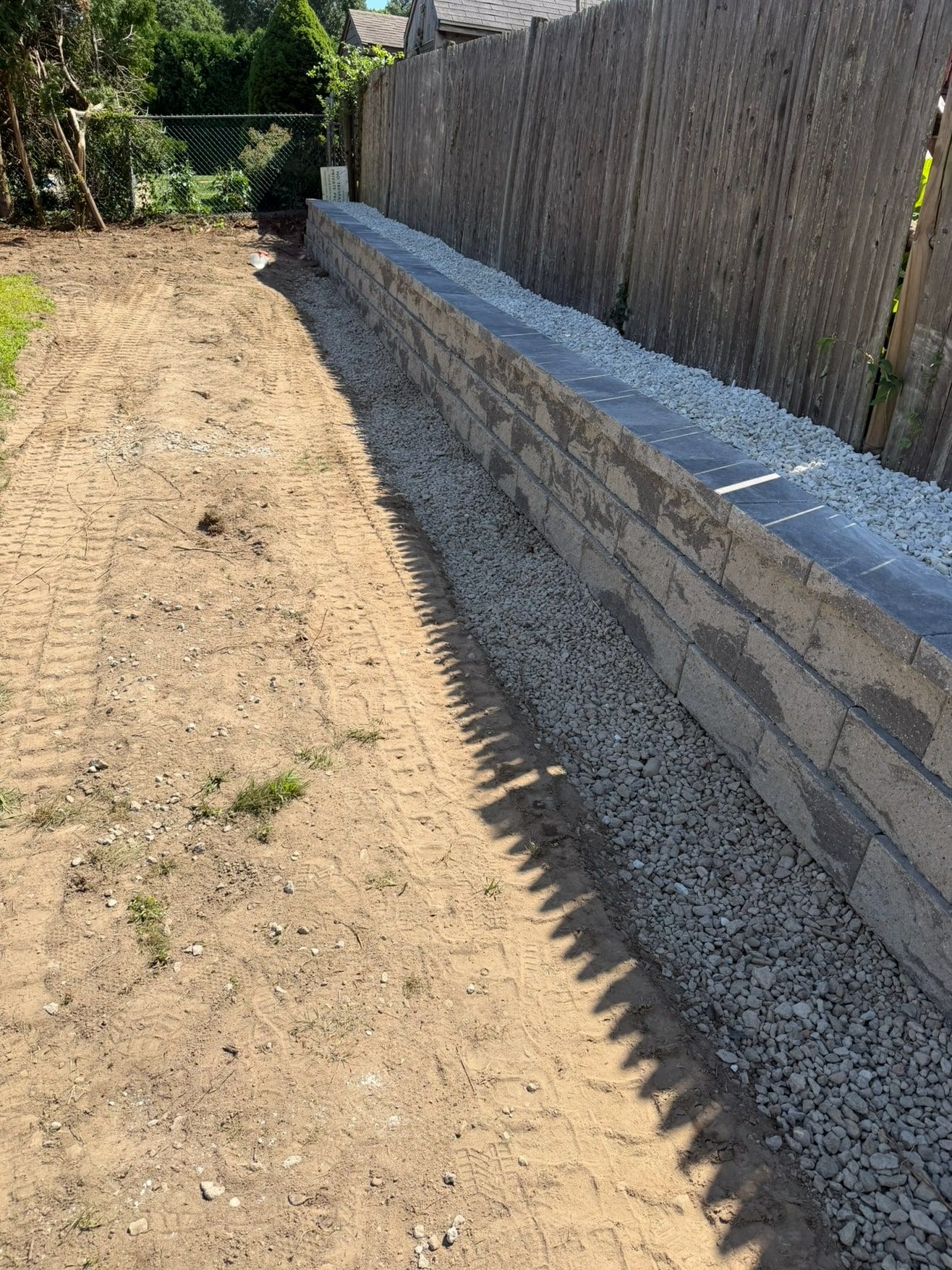 Finished block retaining wall with dark cap stones and crushed-stone drainage along a wooden fence line